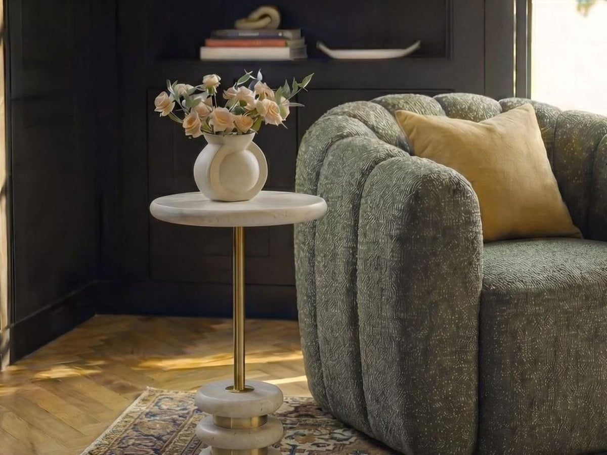 Rémi Travertine Drink Table styled beside a curved gray lounge chair in a dark-paneled interior.