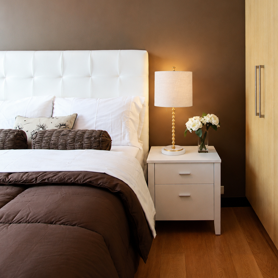 Adèle table lamp with a white linen shade, sculpted gold-finished stem, and round white marble base styled on a white nightstand beside a bed in a warm modern bedroom.