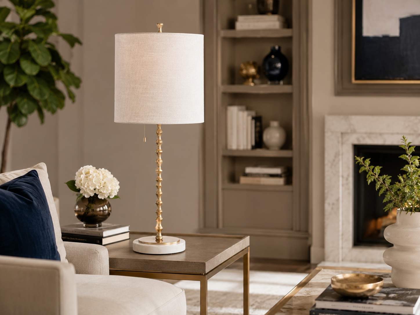 Adèle table lamp with a white linen drum shade, sculpted gold-finished stem, and round white marble base styled on a console against a deep teal wall with layered gold and black décor.