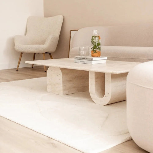 Light travertine coffee table with sculptural U-shaped leg styled in a neutral living room with a beige armchair