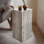 Travertine cube side table with natural vein-cut texture styled beside a gray armchair with a vase and candle