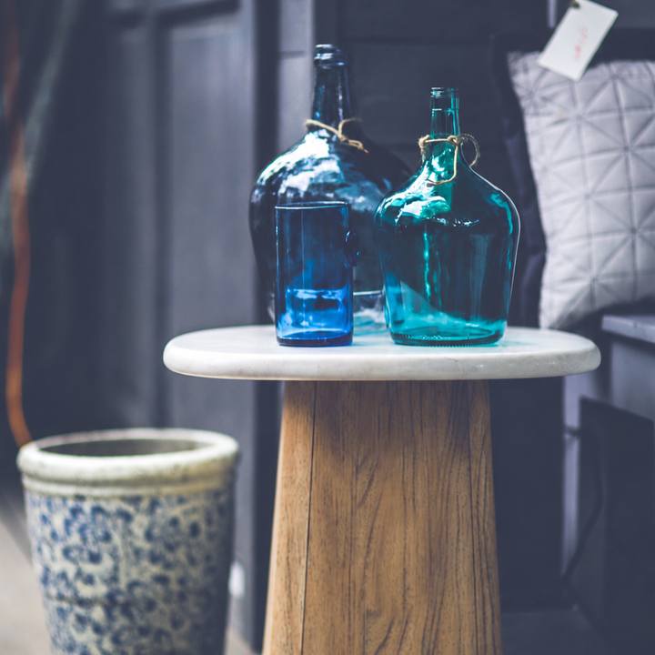 Portico Travertine Accent Table styled with blue glass vessels in a moody interior, contrasting the pale stone top with the warm wood pedestal.