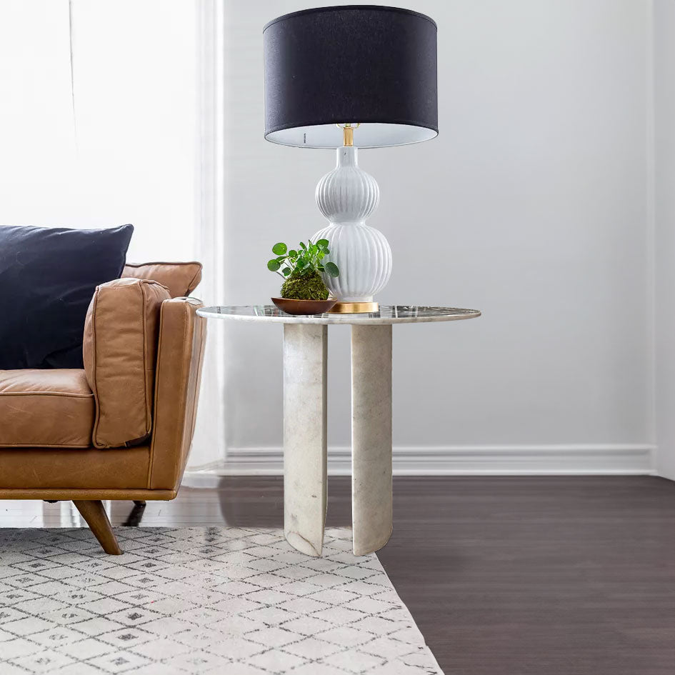 Round marble side table with twin curved legs styled with a table lamp beside a brown leather sofa