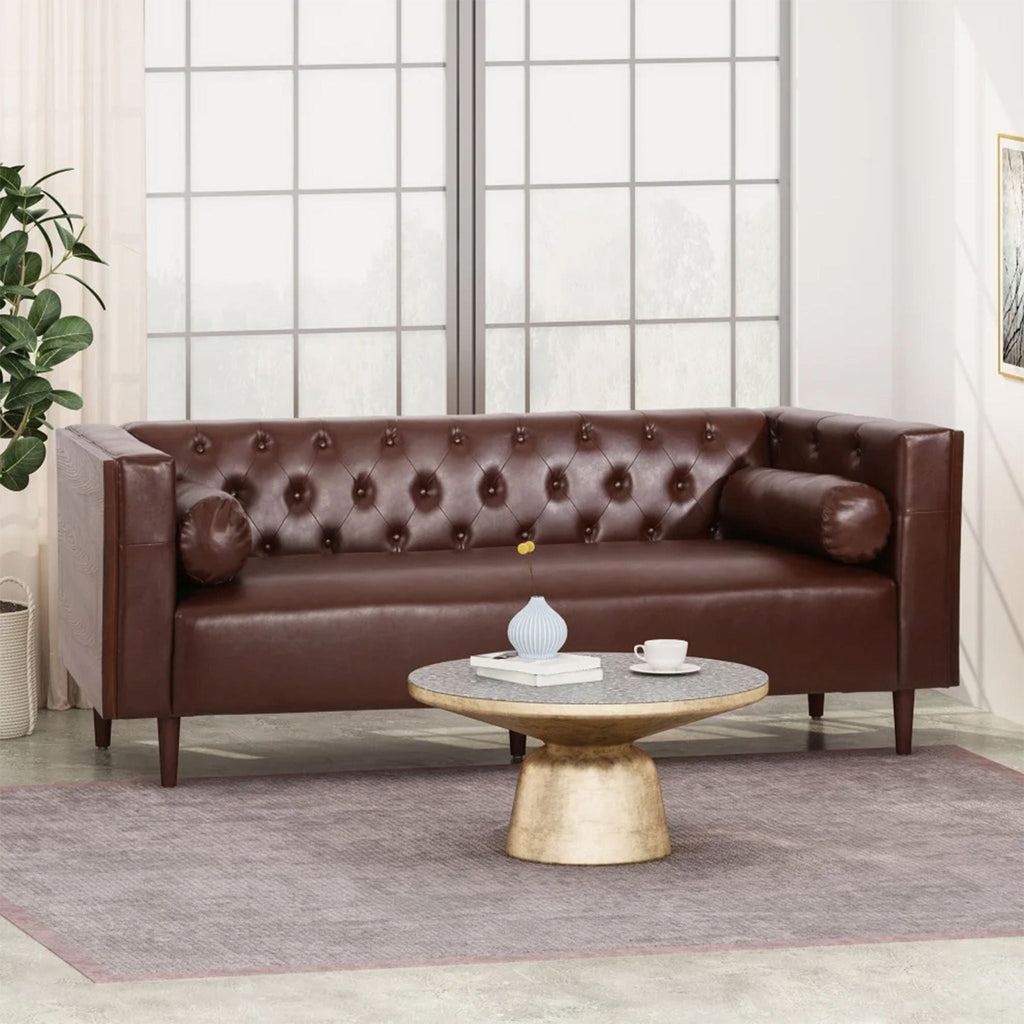 Luxury 90-inch tuxedo sofa from the Monolith collection in premium dark brown leather, featuring an interior grid of deep button tufting. Shown in a styled editorial lounge space behind a round gold-based coffee table.
