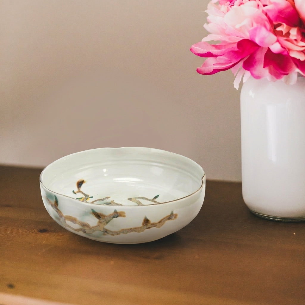 Nadine Bowl styled on a warm wood surface beside a white vase filled with vivid pink flowers, highlighting its softly irregular shape and hand-painted branch motif.