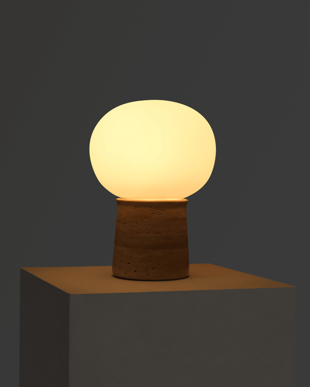 The Noémie Table Lamp illuminated, showing the warm glow of its glass shade against the textured travertine base in a dark setting.