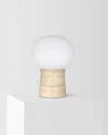 The Noémie Table Lamp with a frosted-glass orb shade resting on a weathered travertine base, shown unlit against a light background.