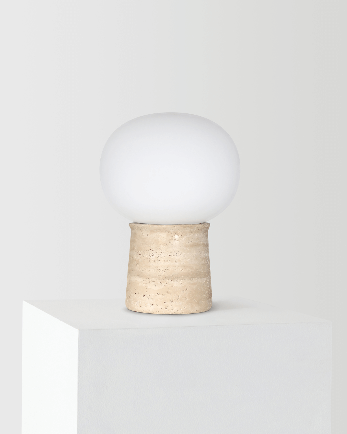 The Noémie Table Lamp with a frosted-glass orb shade resting on a weathered travertine base, shown unlit against a light background.