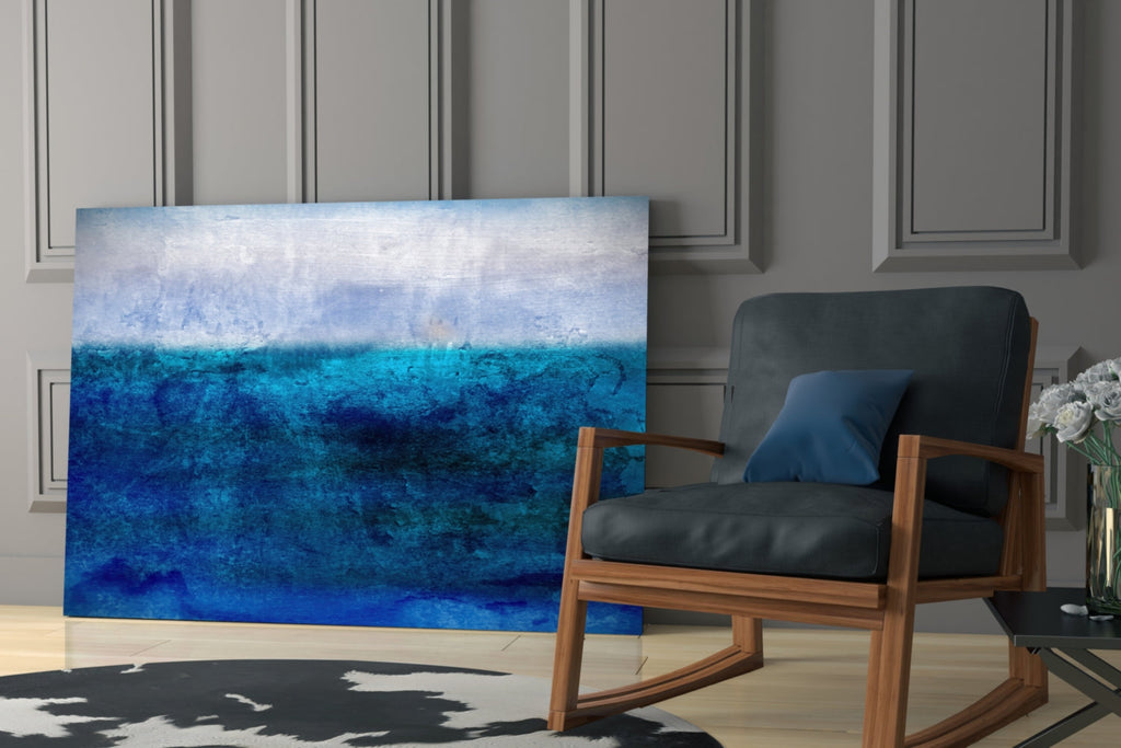 Large horizontal blue abstract glass wall art leaning against a wall beside a wooden armchair