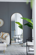 Brushed gold arched floor mirror leaning against a charcoal wall beside a cream sofa and greenery