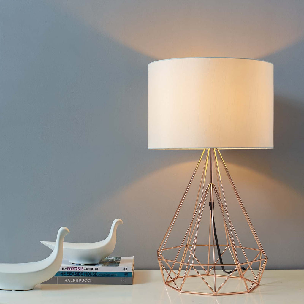 Rose gold geometric table lamp illuminated beside stacked books and ceramic bird figurines on a console table