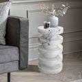 Stacked white marble side table styled with a vase and candle beside a gray velvet sofa