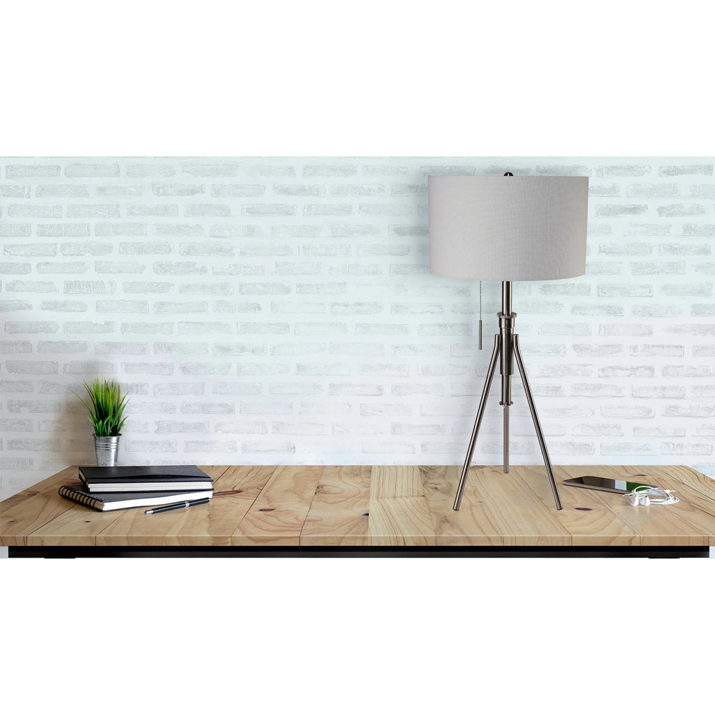 Brushed nickel adjustable tripod lamp styled on a wood table against a whitewashed brick wall