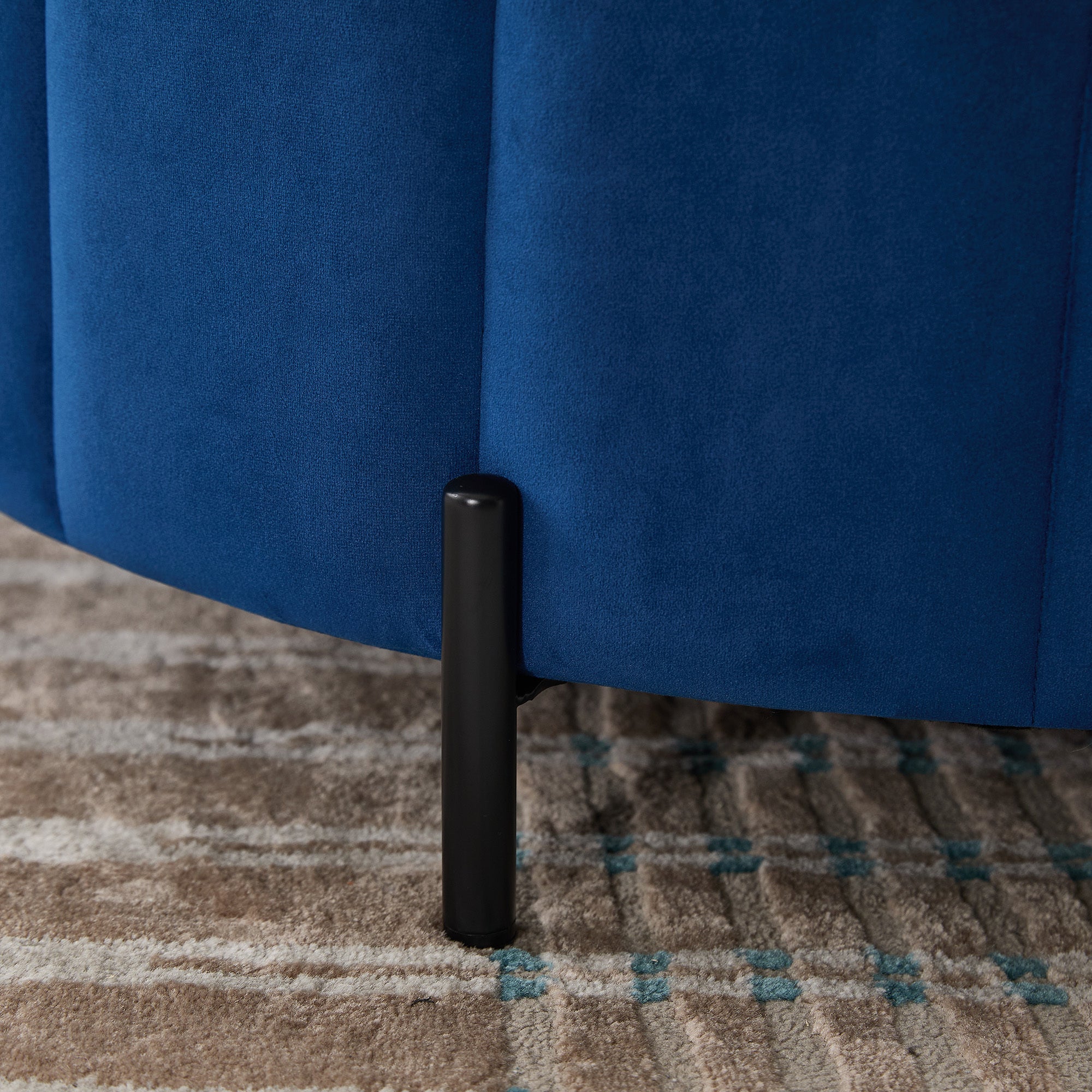 Close-up of the Charlotte Ottoman's slender black adjustable metal leg against navy channel-tufted velvet