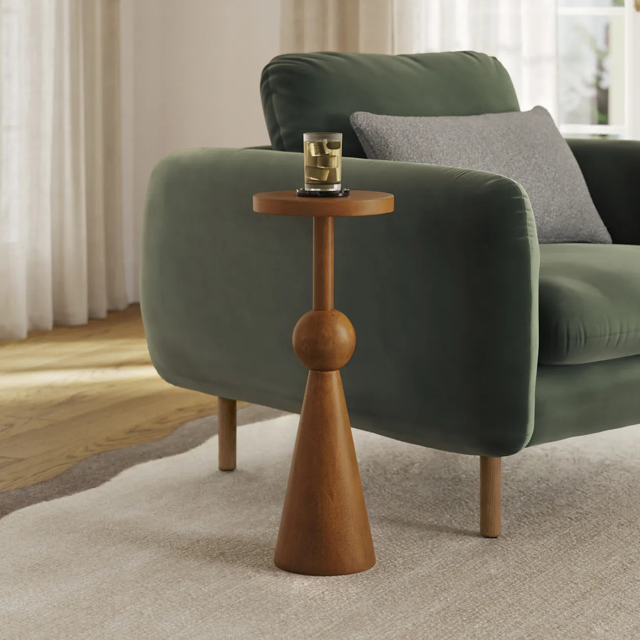 Walnut sculptural drinks table holding a glass beside a green velvet armchair