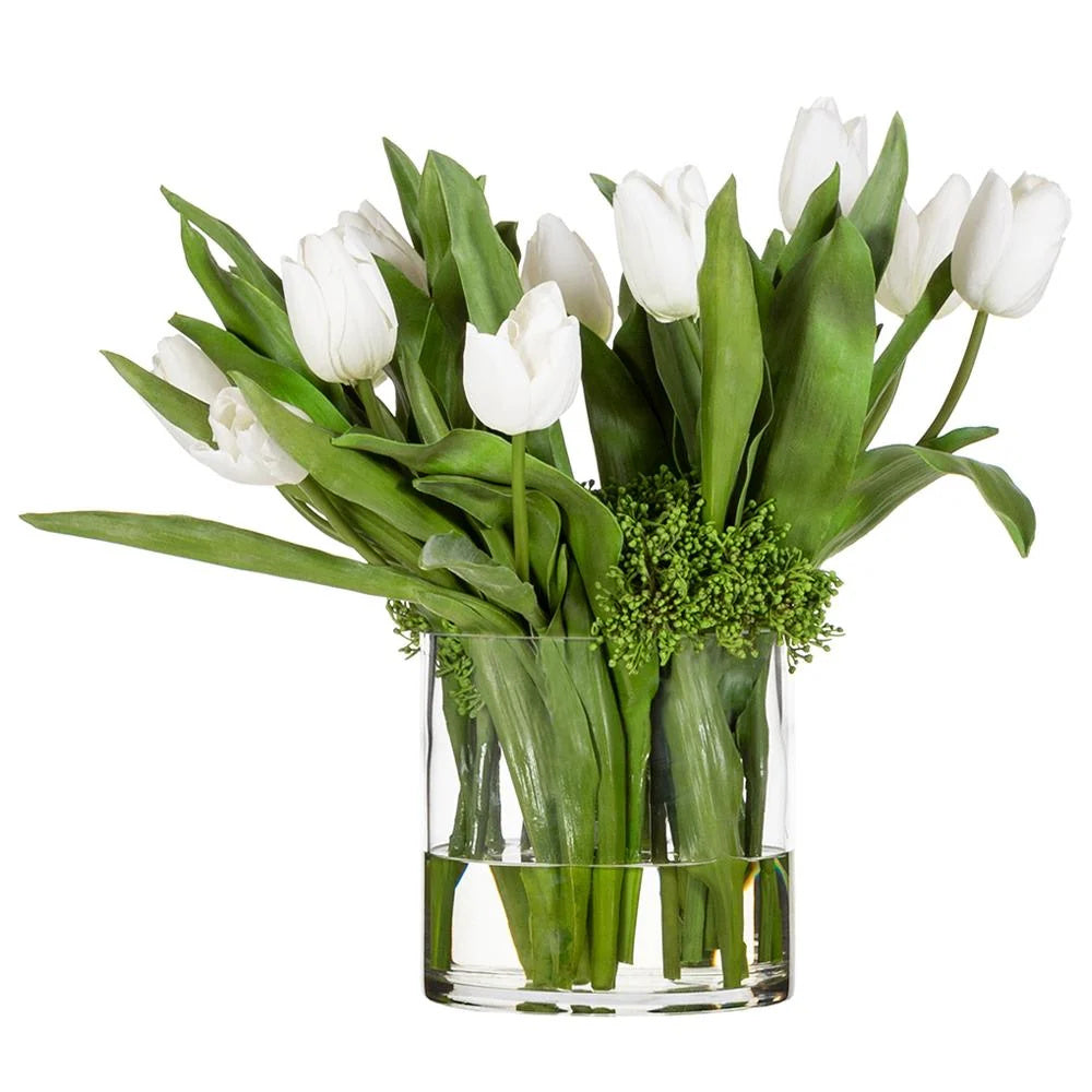 Studio view of the Inès Tulip Arrangement showing white artificial tulips, long green leaves, compact green filler, and a clear cylindrical glass vase