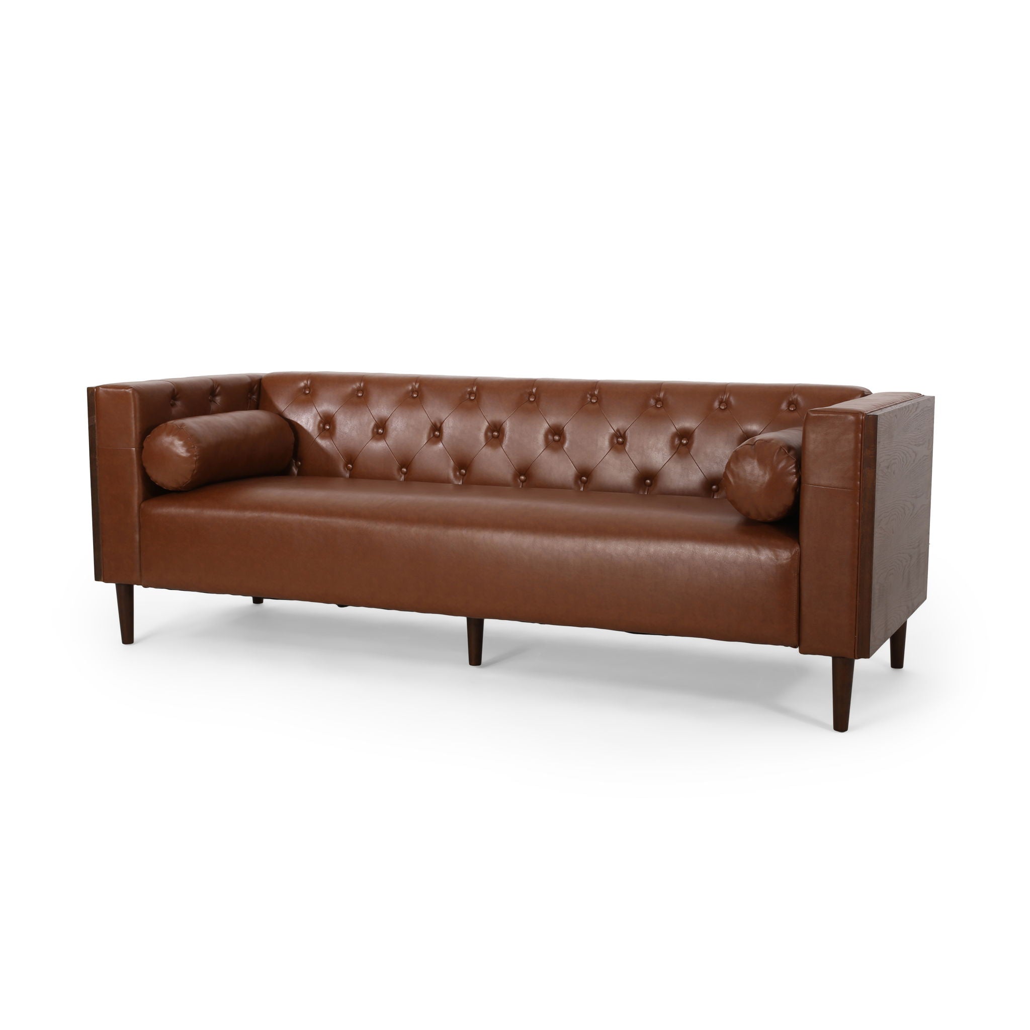 Three-quarter studio angle of the Monolith tuxedo sofa wrapped in rich caramel brown leather, highlighting the deep internal diamond button tufting and matching cylindrical bolster pillows.