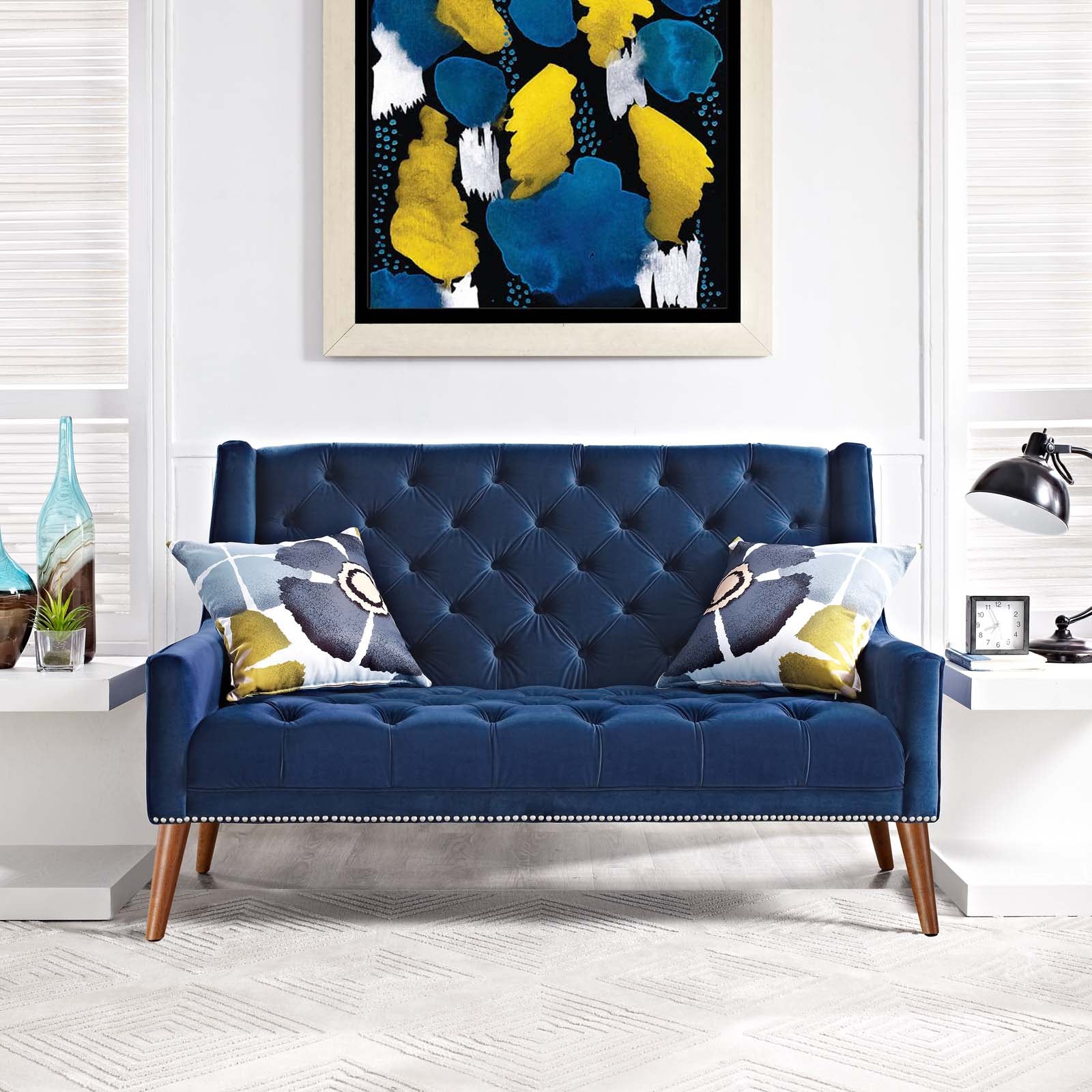 Editorial living room view of the Peruse velvet loveseat in navy blue, styled with abstract floral pillows on a white geometric rug beneath a bold yellow and blue canvas painting.