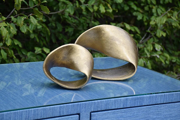 Large and small Ruben sculptures styled together on a blue console, their warm metallic loops contrasting with the cool surface and green foliage behind.