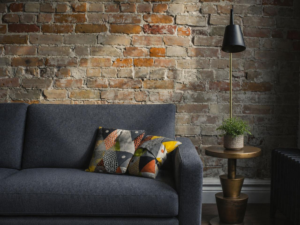Victoire Side Table styled beside a charcoal sofa against an exposed brick wall with a small green plant on the round top.