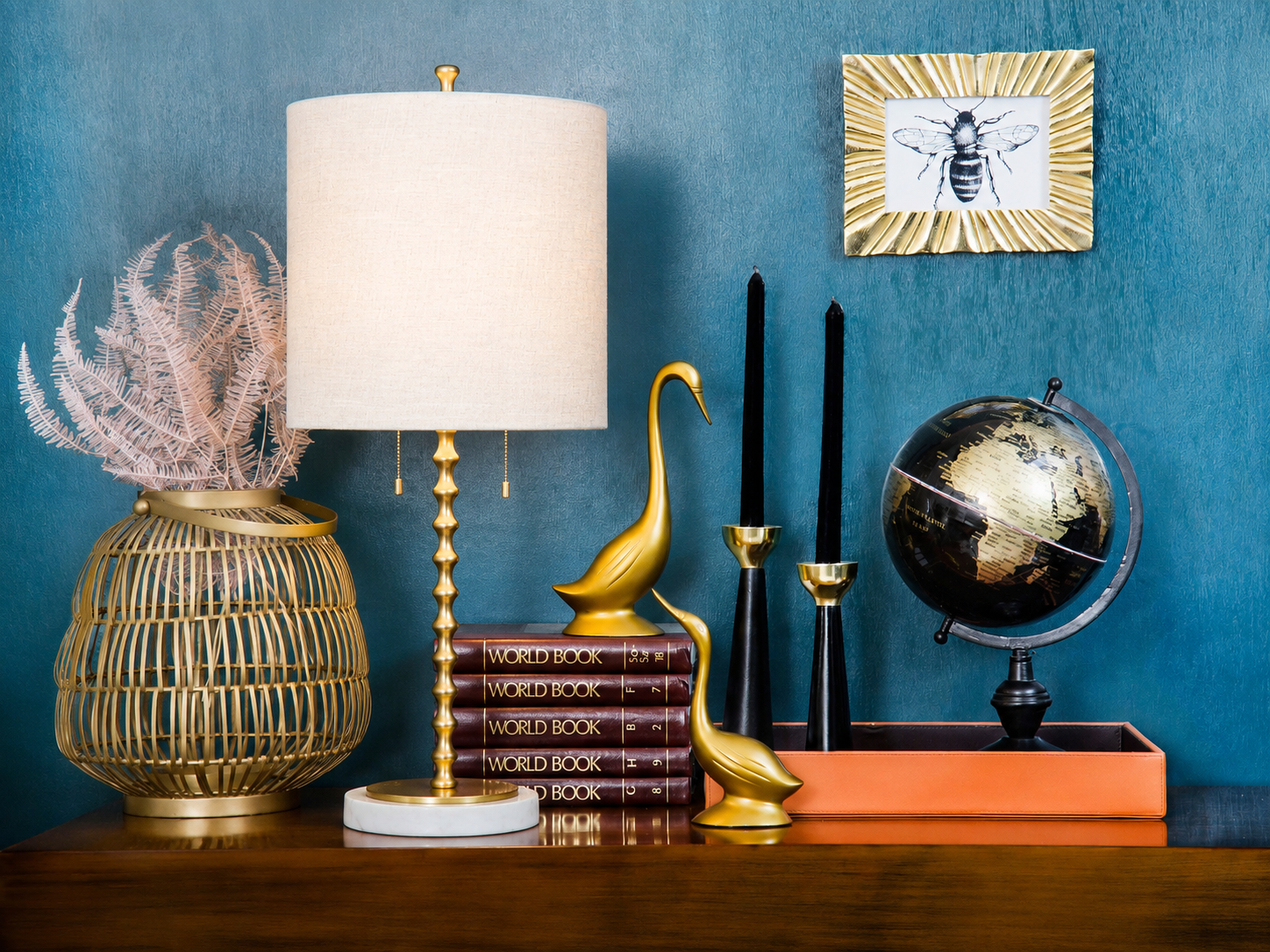 Adèle table lamp with a white linen drum shade, sculpted gold-finished stem, and round white marble base styled on a console table with layered decorative objects in a richly colored interior.